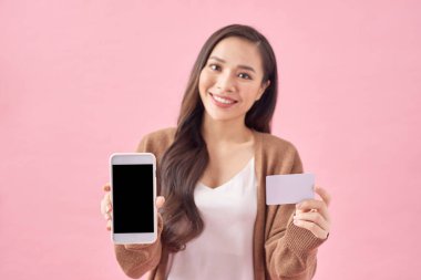 Pembe arka planda soyutlanmış mutlu Asyalı genç bayan görüntüsü. Telefon kullanırken kameraya bakmak ve bankamatik kartı tutmak.