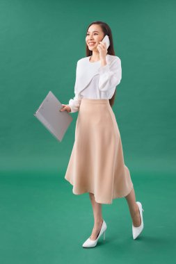 Turkuaz arka planda yalnız poz veren mutlu genç kadın cep telefonuyla konuşan klasörü tutuyor..