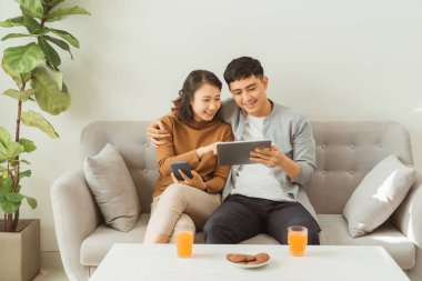 Genç bir çift koltukta tablet ve cep telefonu kullanırken evde keyfine bakıyor.