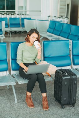 Genç iş kadını havaalanında otururken dizüstü bilgisayar kullanıyor ve kahve içiyor..