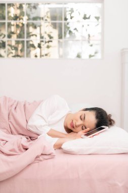 Rahat, genç, güzel bir kadın boş ekran cep telefonuyla yatakta uyuyor.