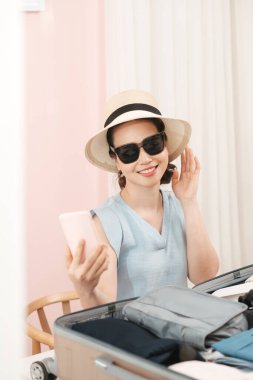 Güzel kadın gezgin selfie çekiyor ve moda şapkasını seçiyor. Evde tatil tatili için bavula giysiler koyuyor.