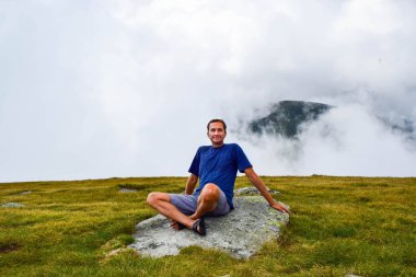 Adam Transalpina yolu yakınlarındaki sis ve alçak bulutlarla çevrili bir taşın üzerinde oturuyor. Burası Romanya 'nın en güzel dağlık yolları ve en yüksek asfalt yollarından biri..