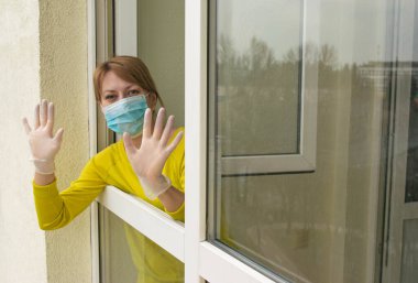 Koruyucu maskeli ve eldivenli kadın, kendini izole etme ve salgın ve karantina süresince evde kalma tavsiyelerine uyarak evinin açık penceresinde duruyor..