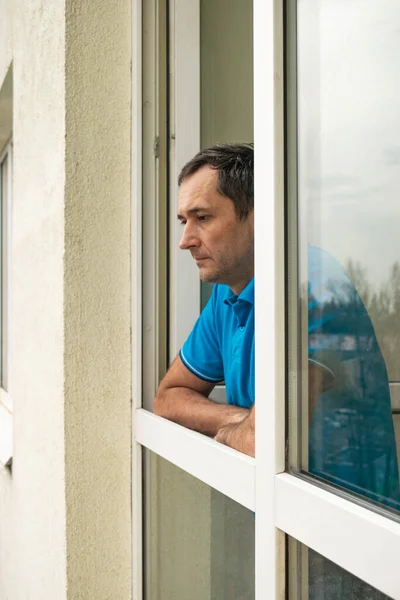 The pensive man looks at the street, standing at the open window of his apartment, following the recommendations to stay at home during the epidemic and quarantine.