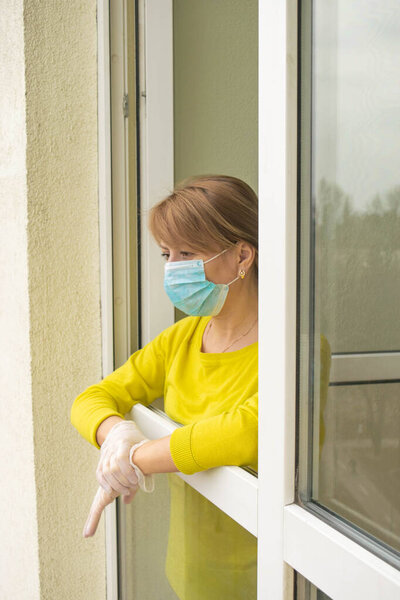 The woman in a protective mask and gloves stands at the open window of her apartment, following the recommendations of self-isolation and staying home during the epidemic and quarantine.
