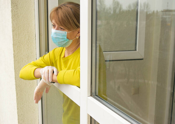 The woman in a protective mask and gloves stands at the open window of her apartment, following the recommendations of self-isolation and staying home during the epidemic and quarantine.
