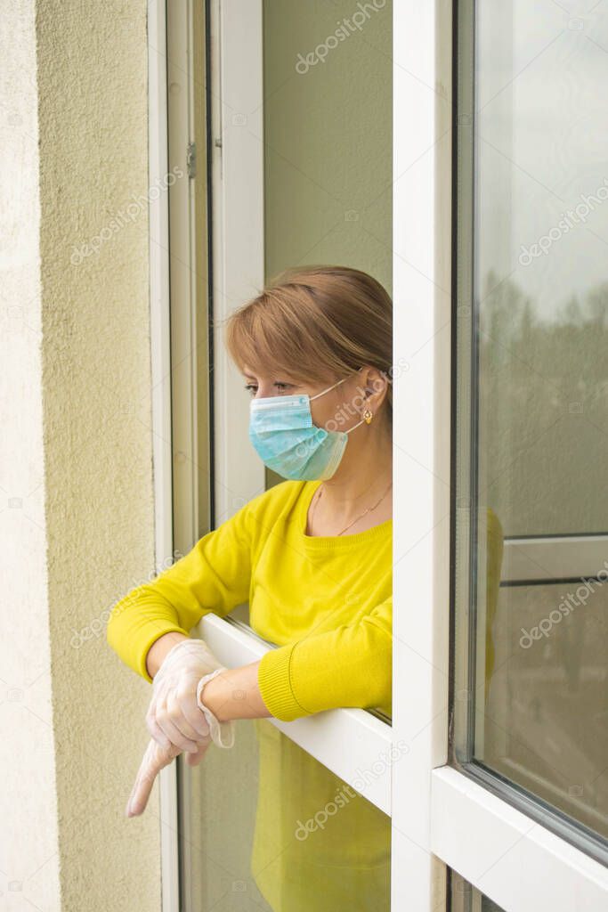 The woman in a protective mask and gloves stands at the open window of her apartment, following the recommendations of self-isolation and staying home during the epidemic and quarantine.
