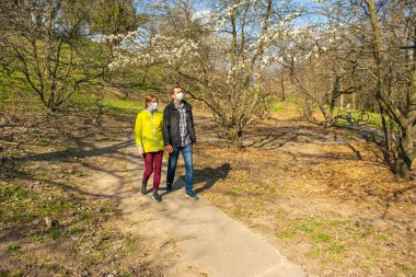 Koruyucu sağlık maskeli bir erkek ve kadın çifti güneşli bir ilkbahar öğleden sonrasında parkta yürürler. Coronavirus salgını ve salgını sırasında güvenlik önlemleri.