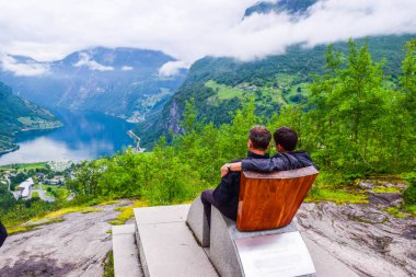Flydalsjuvet Viewpoint 'te Queen Sony Chair' de oturan erkek turistler. Geirangerfjord 'un sonunda yer alan Geiranger küçük köyünün yaz manzarası. Norveç.