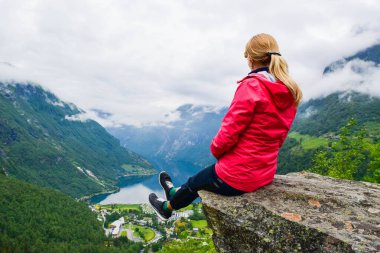 Flydalsjuvet Viewpoint yakınlarındaki uçurumun kenarında oturan kadın turist. Geirangerfjord 'un sonunda yer alan Geiranger küçük köyünün yaz manzarası. Norveç.