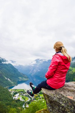 Flydalsjuvet Viewpoint yakınlarındaki uçurumun kenarında oturan kadın turist. Geirangerfjord 'un sonunda yer alan Geiranger küçük köyünün yaz manzarası. Norveç.