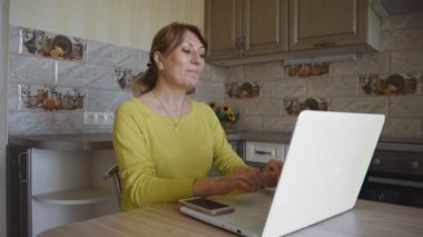 Genç, beyaz, gülümseyen bir kadın internet üzerinden iletişim kurarken elini sallıyor. Evde, mutfakta bir masada oturuyor. Sosyal ağlar ve ev eğlencesi.