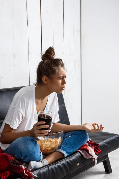 Girl eating chips, drinking soda, watching tv, sitting at sofa.