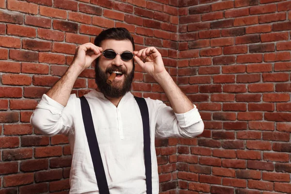 Young handsome man laughing holding cigar over brick background.