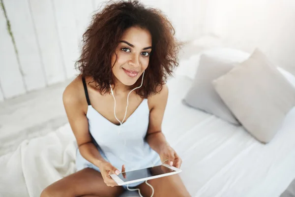 Beautiful african girl in sleepwear smiling looking at camera listening to music in headphones sitting on bed.