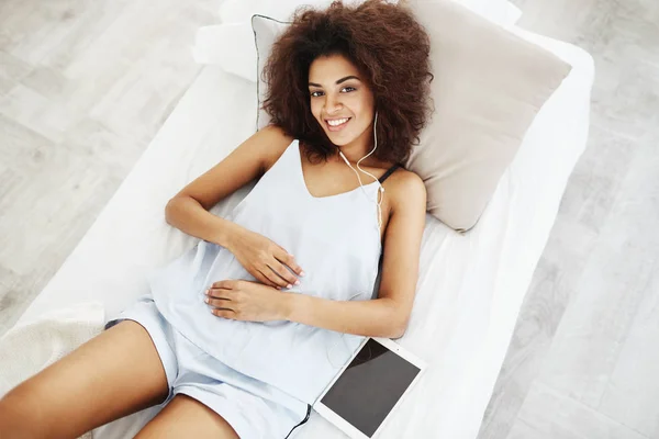 Beautiful african girl in sleepwear smiling looking at camera listening to music in headphones lying on bed. From above.