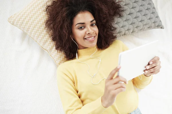 Beautiful african girl in headphones holding tablet lying on bed smiling looking at camera. From above.