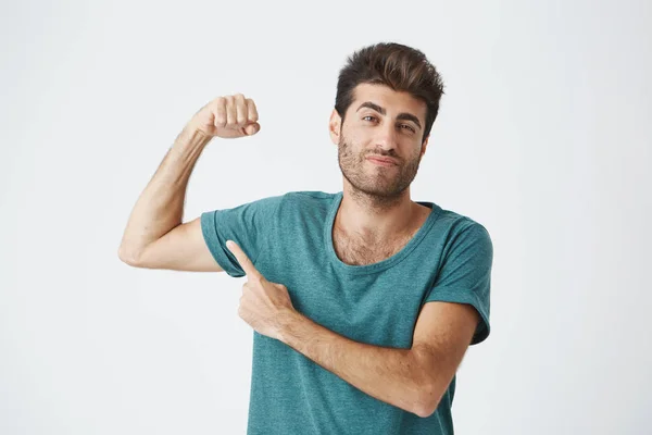 Man with stylish hairstyle and beard frowning his eyebrow, demonstrating how strong he is. Fit guy pointing with his finger at his biceps. Macho man showing himself.