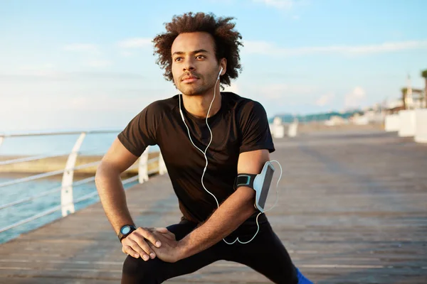 Fit male Afro-American jogger with bushy hairstyle warming-up his muscles before running. Man athlete in black sportswear stretching legs with stretch exercise on wooden pier with white earphones on.