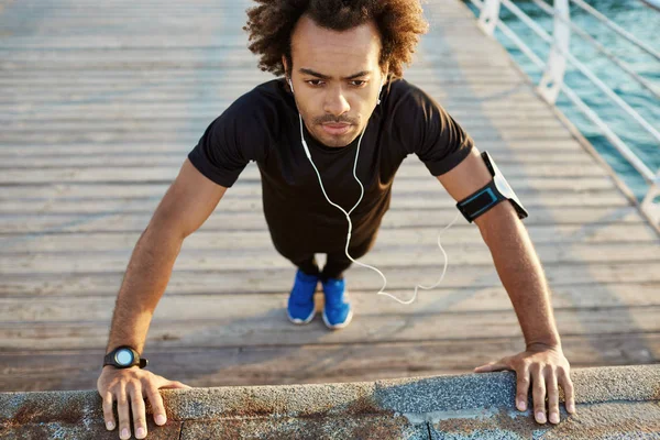 Dark-skinned man runner in black sportswear standing in plank position warming up before cardio workout on the pier in the morning. Afro-american male athlete wearing black sport suite and blue