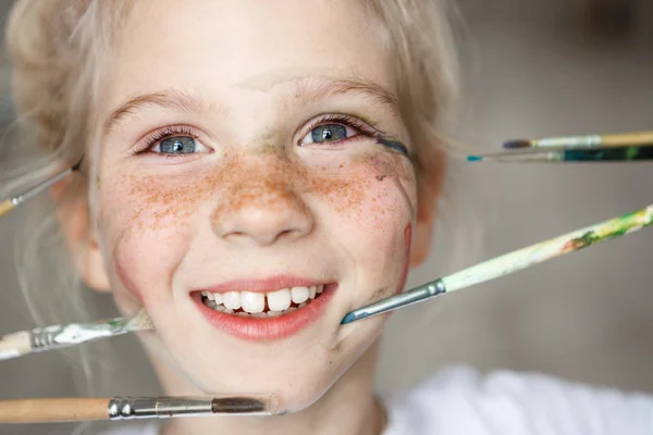Portrait of playful blonde European girl with freckles and paint on her face, playing with brushes. Blonde pretty cheerful girl smiling with teeth.