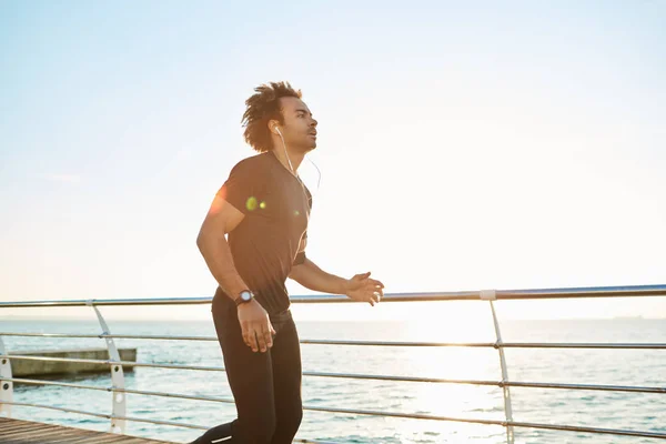 Fitness and sports. Stylish dark-skinned man runner in sportswear doing cardio workout on beach in the morning. Male athlete wearing black sport suite running on the pier listening music through white