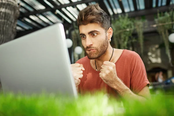 Shot of worrying focused man with stylish hairstyle dressed casually closely looking at the screen of his notebook, watching online translation of match, clenching his fists in nerves.