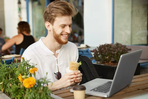 Portrait of mature red-haired guy with beard in casual white shirt resting from work, eating sandwich for lunch and looking funny video in network.