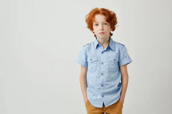 Beautiful little boy with ginger hair and freckles holding hands in pockets looking in camera having serious but unconfident look.