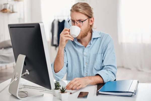 Serious concentrated on work office worker with fair hair, beard in casual outfit and glasses, prepares report, uses keyboard, drinks coffee, works during lunch break, sits against office interior.