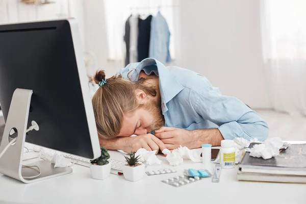 Tired sick fair-haired man, suffering from headache and high temperature, keeping head on hands, sitting in front of computer screen, covering face. Ill office worker surrounded by pills and drugs