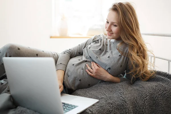 Spending pregnancy period with joy. Beautiful blonde young pregnant woman in comfy home clothes lying on bed, looking for good movie to watch on laptop, relaxing, holding hand on her belly.