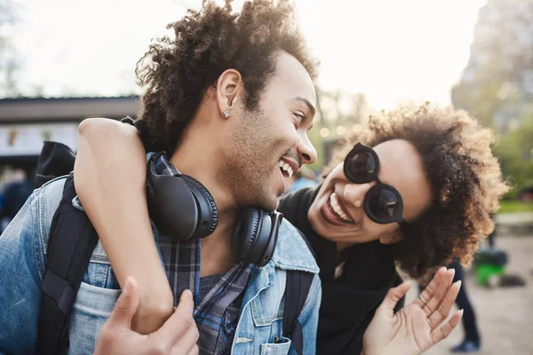 Close-up portrait of cheerful dark-skinned woman hugging boyfriend from back while walking in park and talking, smiling at him. Two friends celebrate their win in university contest