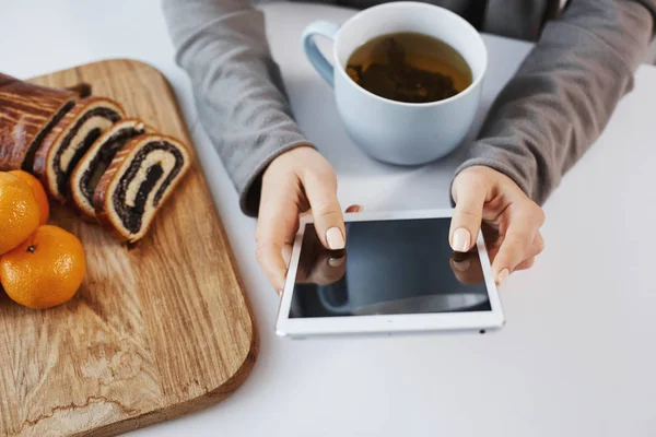 Close-up shot of woman hands holding digital tablet. Girl enjoys weekends in calm and cozy atmosphere, drinking cup of tea and eating tangerines with rolled cake. Businesswoman always stays in touch