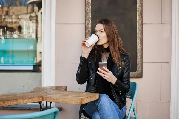 Wild city life consumes lots of energy. Attractive thoughtful and stylish female tourist, sitting cafe patio and drinking coffee, looking aside, thinking about plans for night, holding smartphone