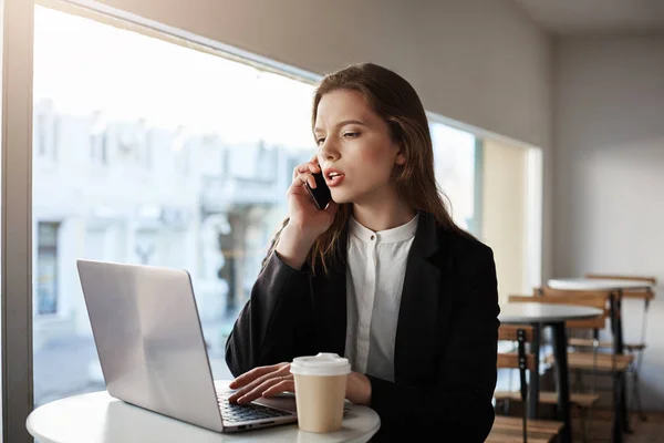 Calling you to inform about stage of work. Indoor shot of serious and focused successful businesswoman in trendy outfit sitting in cafe, drinking coffee, typing in laptop and talking on smartphone