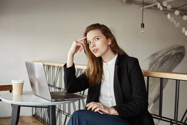 Girl searches inspiration in coffee shop. Indoor portrait of gorgeous slender woman in stylish outfit sitting in cafe, leaning on table while thinking about new concept for company, working in laptop