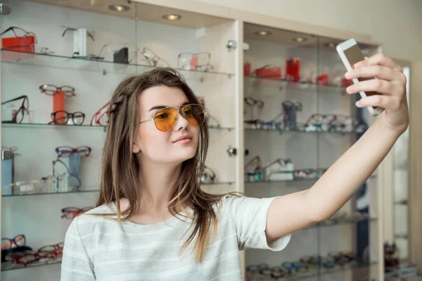 Attractive woman went on shopping alone, making selfie while trying on new stylish sunglasses in optician shop, sending photo to friend so she will say her opinion. Girl bought trendy eyewear