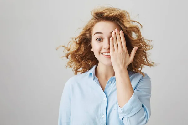 Indoor portrait of excited beautiful caucasian female student with short curly hair covering half of face with palm and smiling, expressing astonishment and positive attitude over gray wall