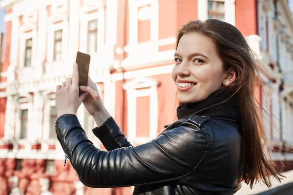 Scenery of town is stunning. Portrait of attractive woman taking photo of museum while being on excursion in foreign city, smiling at camera and talking with husband who travels with her