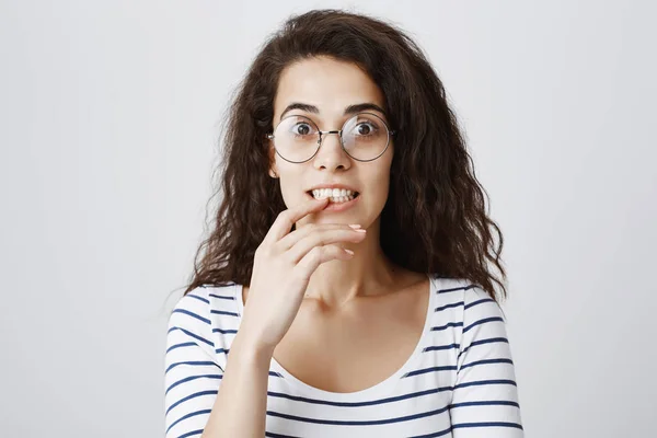 Intrigued girl listens to her friend stories, wanting to hear continuation of story. Smart curly-haired female gazing at camera with curiosity and interest, being nervous, touching teeth with finger