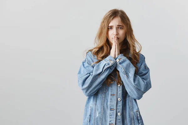 Forgive me for mistakes. Portrait of sad worried attractive female student in denim jacket, holding hands in pray over mouth, frowning, apologizing or begging for help over gray background