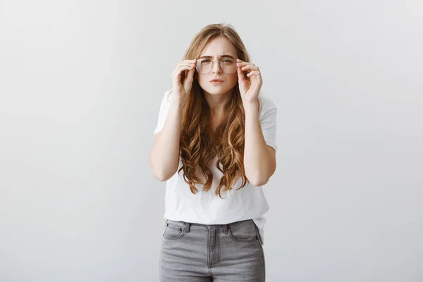 Need to visit optician to buy better glasses. Funny young european female bending towards camera, squinting and holding rim of eyewear while trying to see on long distance but having bad vision