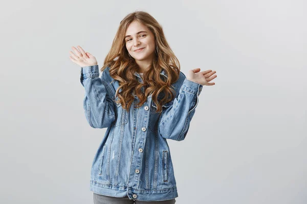 Cute girlfriend bragging about her achievements. Stylish good-looking european woman in denim jacket waving raised palms and smiling with pleased or satisfied expression over gray background