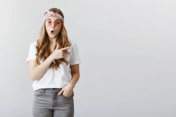 Amazing things happen there. Portrait of wondered excited young caucasian female student in stylish outfit, headband and sunglasses, pointing right and saying wow, being surprised and astonished