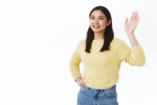 Friendly beautiful asian woman saying hello, smiling happy and waving hand to the left, seeing friend, team leader make goodbye gesture after productive work, feeling pleased and delighted