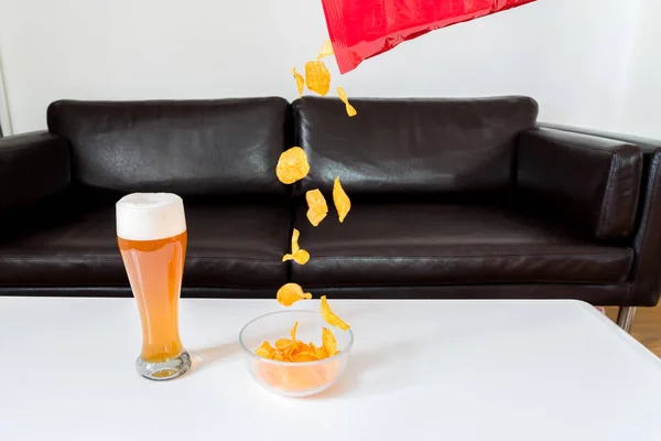 Batatas fritas caindo de um pacote de batatas fritas em uma tigela de vidro com cerveja de trigo na mesa do sofá branco — Fotografia de Stock