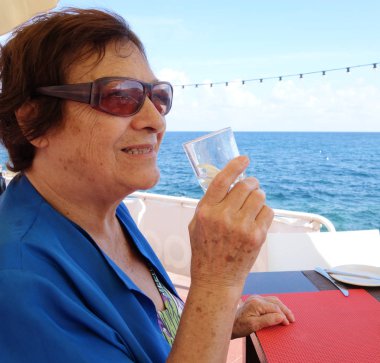 Bardaktan su içen yaşlı bir kadın. Deniz kenarında açık havada çekilmiş.