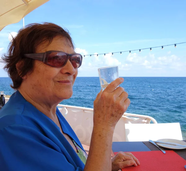 Elderly woman drinking water from a glass. Shot outdoors by the sea